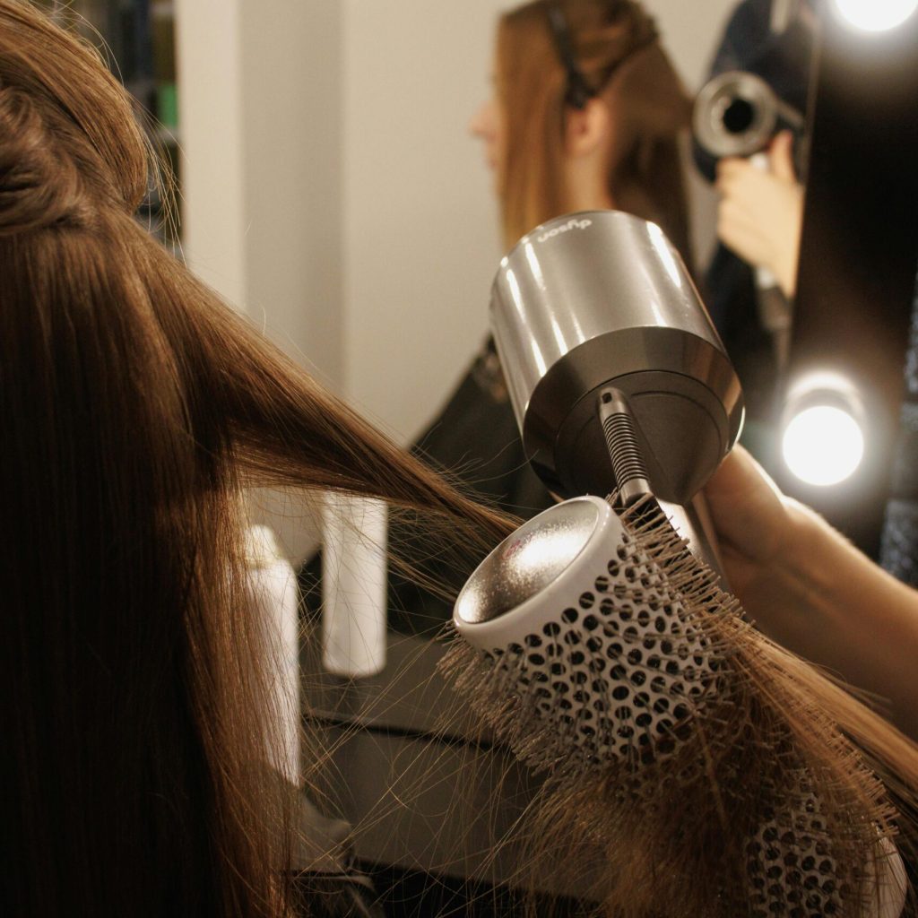 A stylist blow-dries a woman's hair using a brush and hairdryer in a salon.