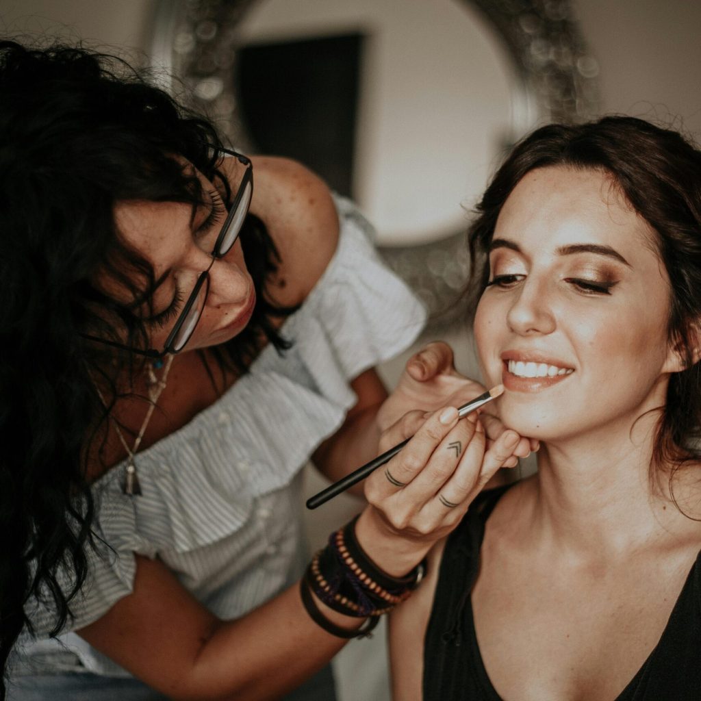 A makeup artist skillfully applies lipstick to a smiling woman in an indoor setting.
