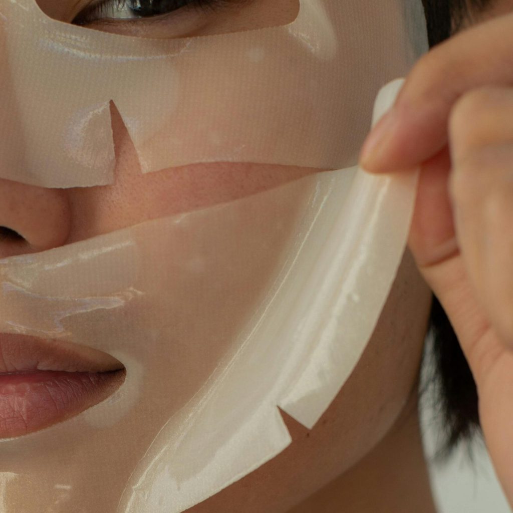 Crop unrecognizable Asian male model looking at camera while taking off moisturizing facial mask during skincare routine in light studio